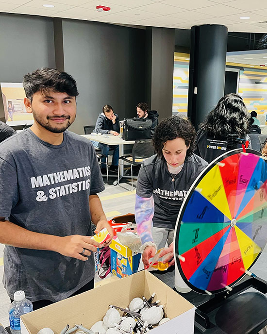 A student in a "Mathematics & Statistics" stands next to a prize wheel.