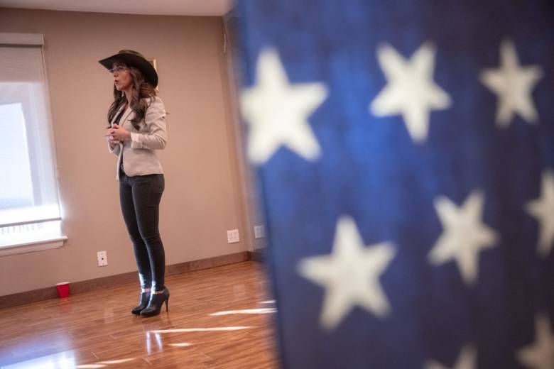 A woman in heels and a cowboy hat talks to a crowd. An American flag covers half the photo.