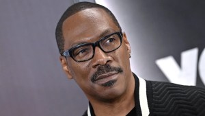 A closeup of a Black man's face. He wears glasses and has a mustache and close-cropped haircut. The man is Eddie Murphy, wearing a black jacket and standing in front of a gray background.