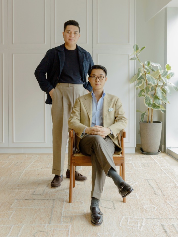 Andy Chong and Buzz Tang, co-founders of The Anthology: Tang is sitting in a chair in front of Chong, in a room with a pale beige patterned carpet, white cupboards on the back wall and a tall pot plant in the corner by a window