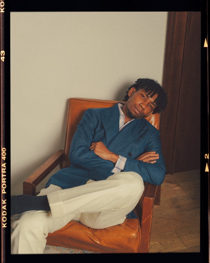 A look from Prologue’s spring/summer 2024 campaign: a man wearing a blue jacket and white trousers, sitting in an orange leather chair with his arms folded