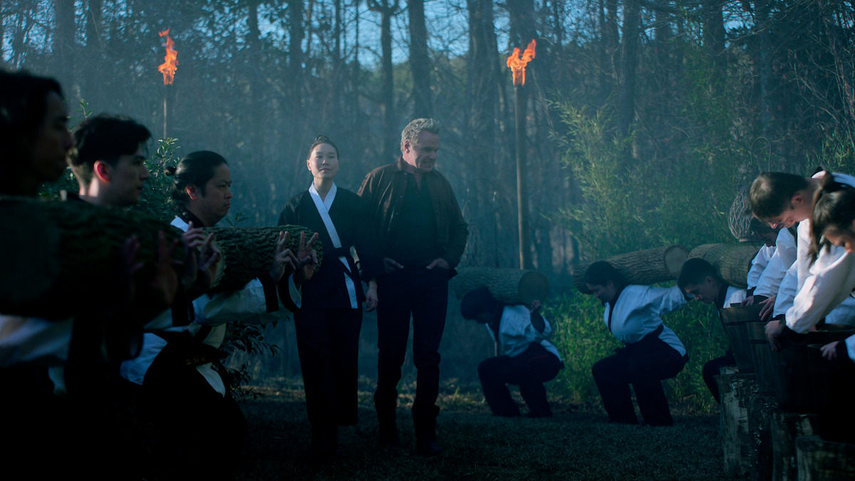 Alicia Hannah-Kim as Kim Da-Eun and Martin Kove as John Kreese walk among students training with heavy longs in Season 6 of ‘Cobra Kai’