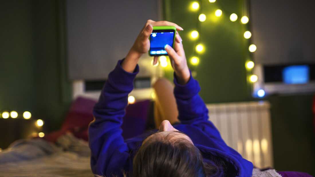 A young girl wearing a long-sleeved blue shirt lays down in her bed, holding her phone. Her bedroom is decorated with strings of white lights. 