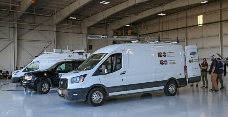 Two white utility vans with logos and rooftop equipment are parked inside a spacious warehouse. Three individuals are conversing near one of the vans.