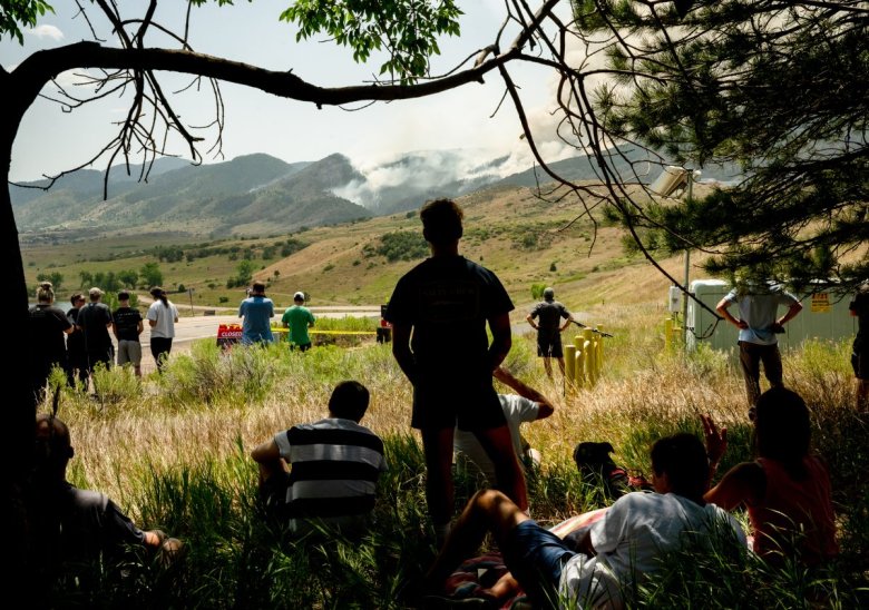 People sitting and standing under trees, watching smoke rise from a distant fire in the mountains on a sunny day.