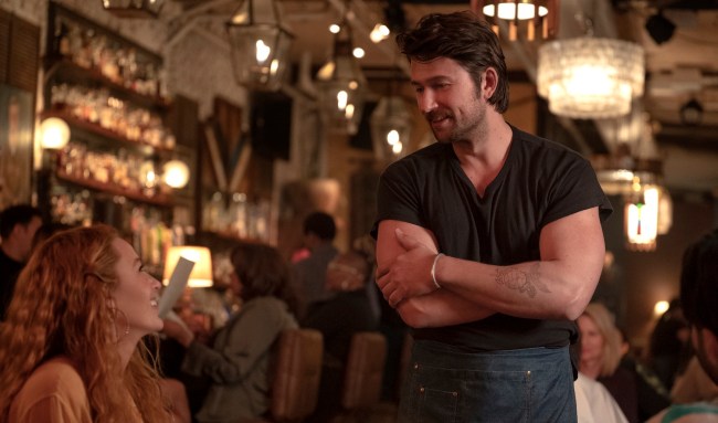 A man in a black t-shirt stands while speaking to a seated woman at a restaurant table; still from 'It Ends With Us'