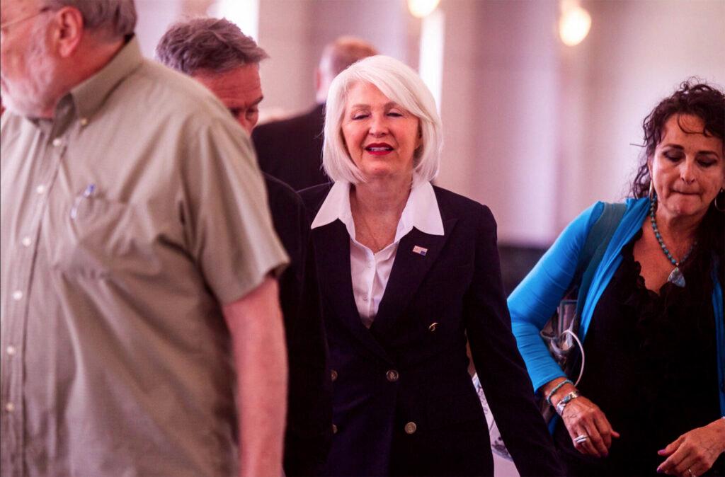 Tina Peters, wearing a blue double breasted blazer with a white shirt, walks down a white hallway outside a courtroom flanked by a supporter wearing a blue shirt.