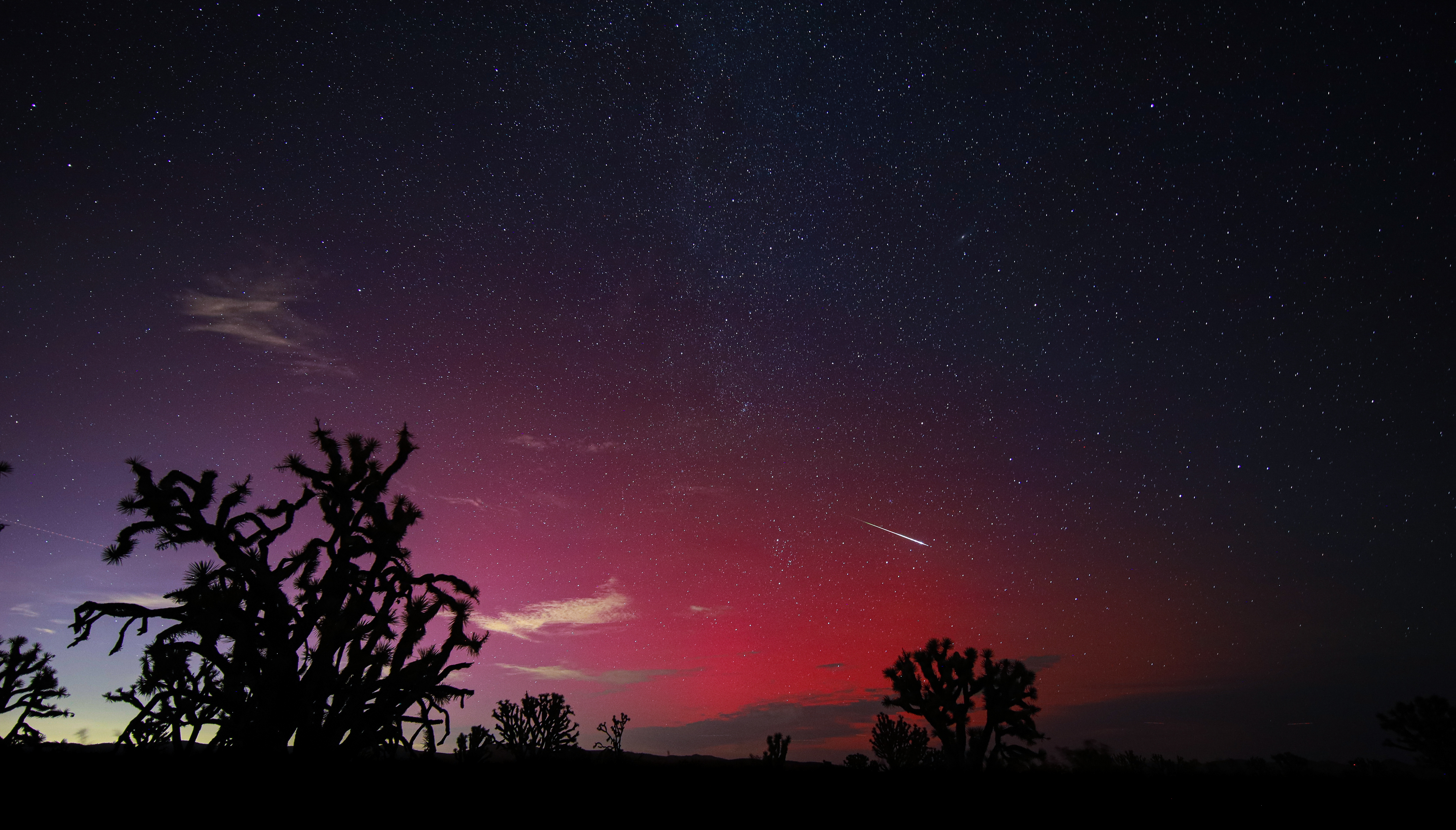 a long white tail of a meteor streaks across the sky which is colored a deep red from the auroras.