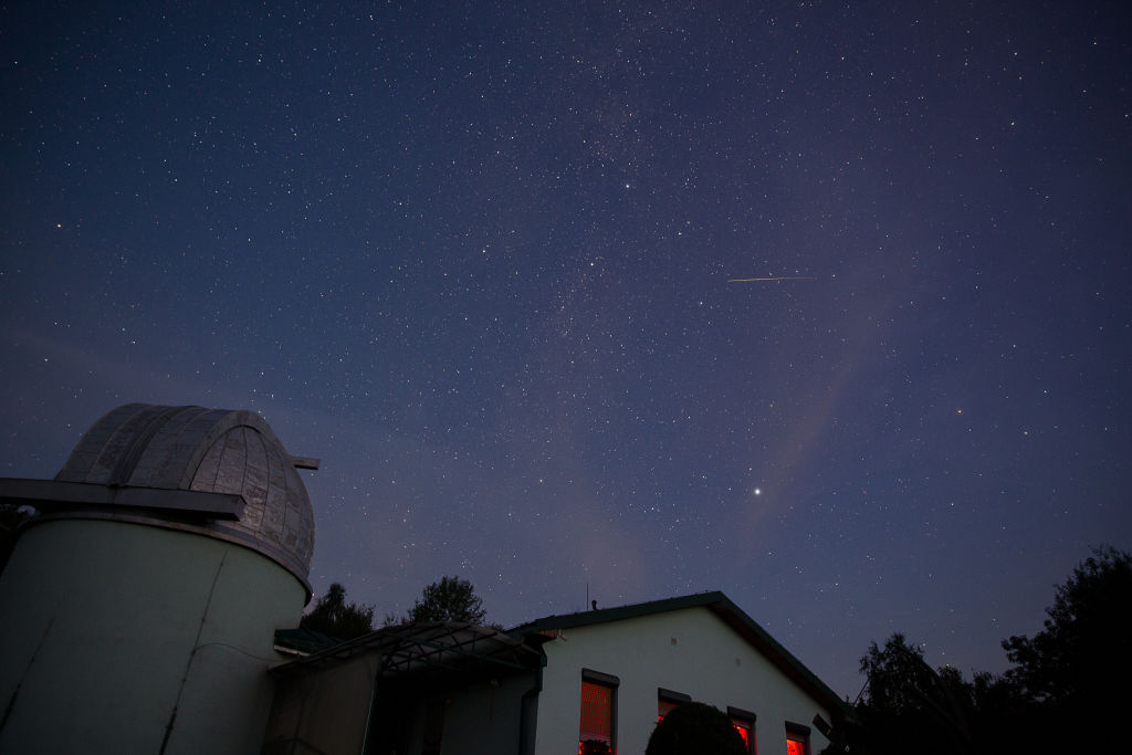 in the foreground is a white building and a small observatory on the left, the sky is filled with stars and a long white streak from a perseid meteor is visible across the sky.