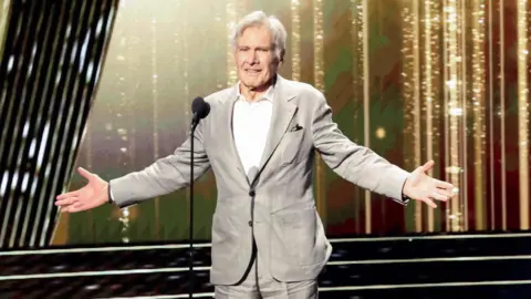 Reuters Harrison Ford gestures as he is honoured at the 2024 Disney Legends Awards during Disney's D23 Expo in Anaheim. Harisson is an 82-year-old man with grey hair. He wears a pale grey suit over a white shirt and stands on stage with his arms outstretched