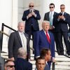 This photo shows former President Donald Trump at Arlington National Cemetery on Monday. He's wearing a blue suit and red tie and is standing outdoors on stairs while surrounded by various officials and staffers in suits.