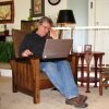 Robert Lang sits on an armchair with a laptop computer resting on his lap. One of his hands is on a computer mouse that sits on one of the armchair's armrests.