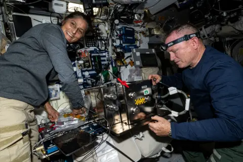 NASA Butch and Suni on ISS - both of them are wearing casual clothing and working on what appears to be complicated equipment with lots of wires and consoles visible. Suni appears to be leaning on one piece of equipment and is smiling, looking over her shoulder at something. Butch has a head torch on and is concentrating on a task.