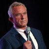 RFK Jr., wearing a suit and holding a microphone, stands against a black backdrop.