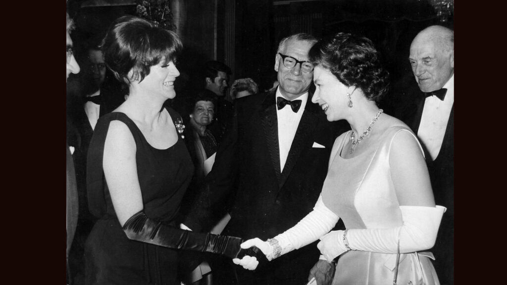 Black and white photo from 1966 of Queen Elizabeth II being presented to Dame Maggie Smith by Sir Laurence Olivier.