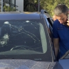 In this photo, a Lebanese police officer wearing a blue shirt and holding a smartphone inspects a car damaged by an exploding pager in Beirut on Tuesday. The car's windshield has multiple cracked areas that resemble spiderwebs.