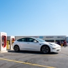 Tesla Superchargers stand in a parking lot in Austin, Texas, on September 16. Four of the five white and red Superchargers in the photo are unoccupied, and a white Tesla sedan is parked in front of one. A strip mall stretches across the background.