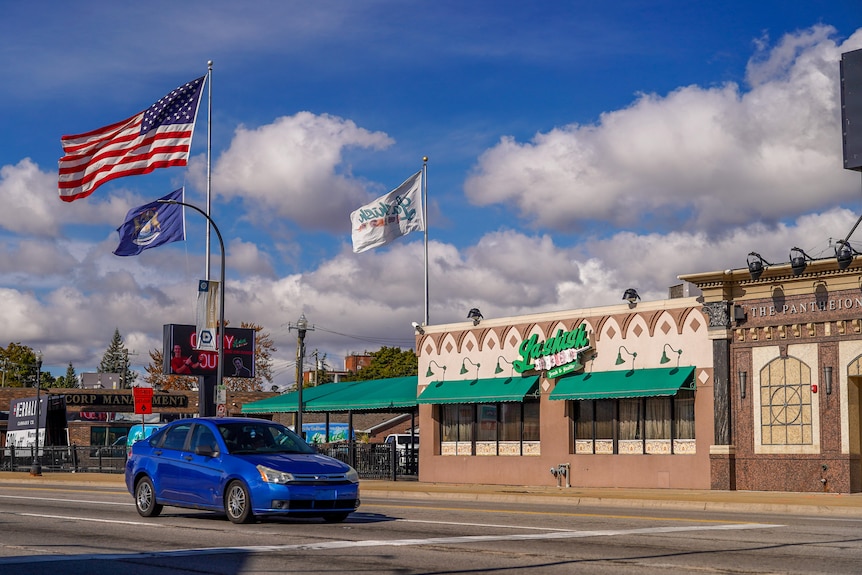 A car drives past a building that says 'Lashish' on its exterior. A large American flag flies from a flagpole.