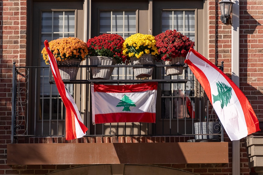 Lebanese flags hang from a balcony of a red-brick building. Colourful flowerpots are also displayed.