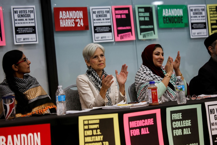 Jill Stein claps while seated at a desk between two other women. Signs say 'abandon Harris' and 'Medicare for all'.