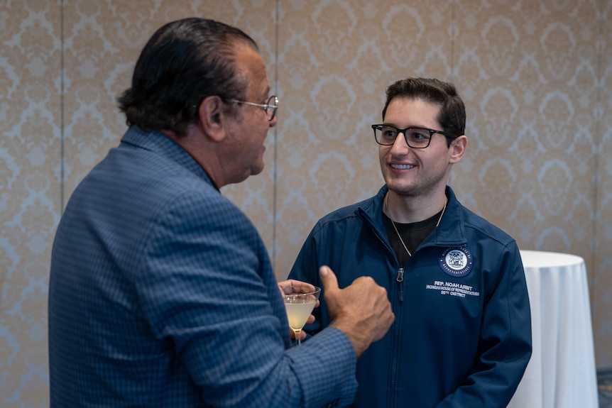 Noah Arbit speaks to another man in front of a wall with ornate wallpaper.