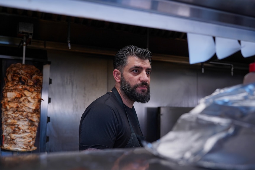 Sam Hammoud stands in a restaurant kitchen. A spit with meat is behind him.