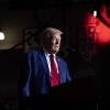 Former President Donald Trump, wearing a suit and a red tie, stands behind a microphone to speak to supporters during a campaign event at Saginaw Valley State University in Michigan. 