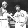 Satchel Paige and Josh Gibson, in their respective Monarchs and Homestead Grays jerseys, talk while holding a ball and a bat.