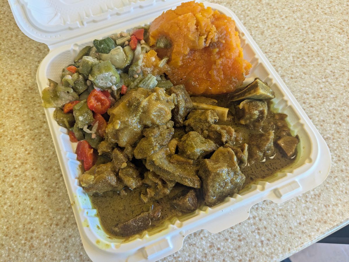 A white plastic container with brown curry, orange yams, and green and red okra and tomatoes.