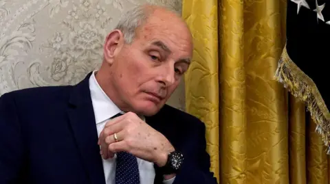 Reuters White House chief of staff John Kelly sits with a gold curtain in the background while wearing a blue suit and tie