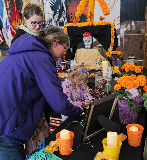 Joan Ribbons, left, her daughter Elyse, and grand daughter JoVi, 4, write a note to their loved one at a community ofrenda at a Dia de los Muertos exhibit at Lansing City Hall Friday, Nov. 1, 2024.