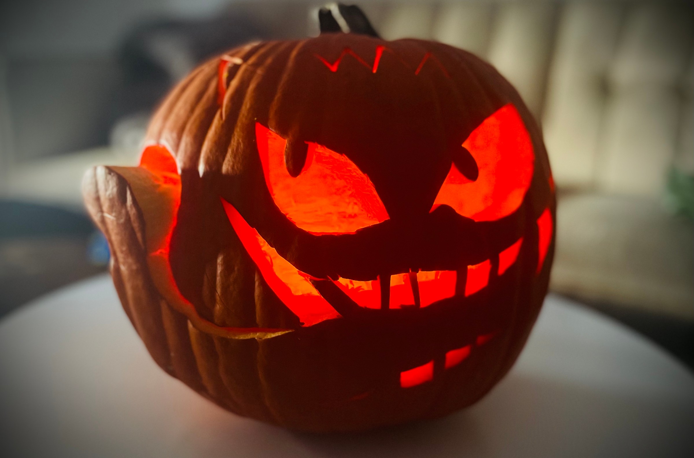 A jack-o-lantern with cat-shaped eyes pointing different directions and large teeth carved into its face. A piece of pumpkin has been carved out like a tongue wrapping around.