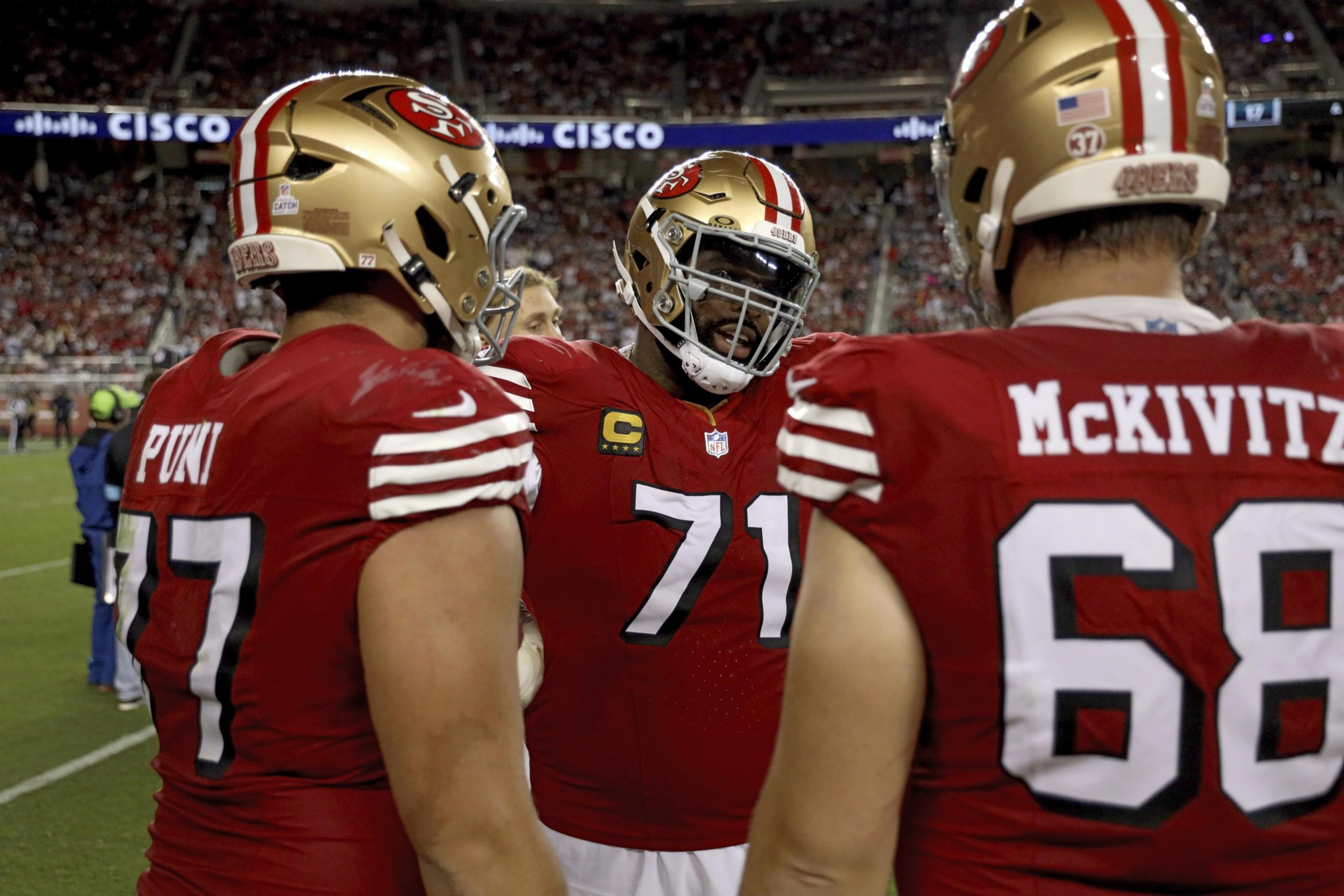 Three football players wearing red 49ers uniforms and helmets stand in a huddle on the field, with a crowded stadium in the background.