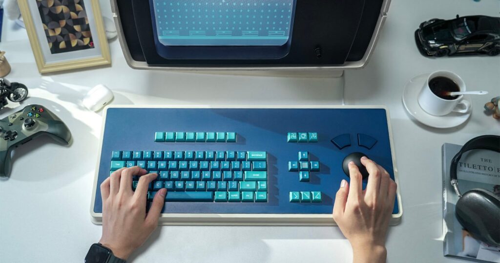 A person types on a retro-style blue keyboard with a large trackball, in front of a vintage monitor. The desk holds a cup of coffee, a book, a toy car, a framed picture, headphones, and a game controller.
