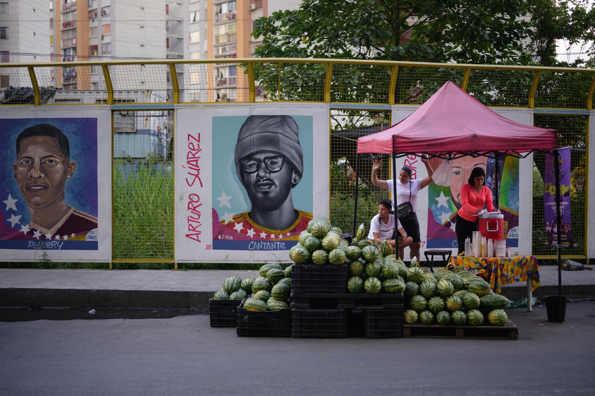 Posters depicting individual people line a fence near a street vendor selling watermelons