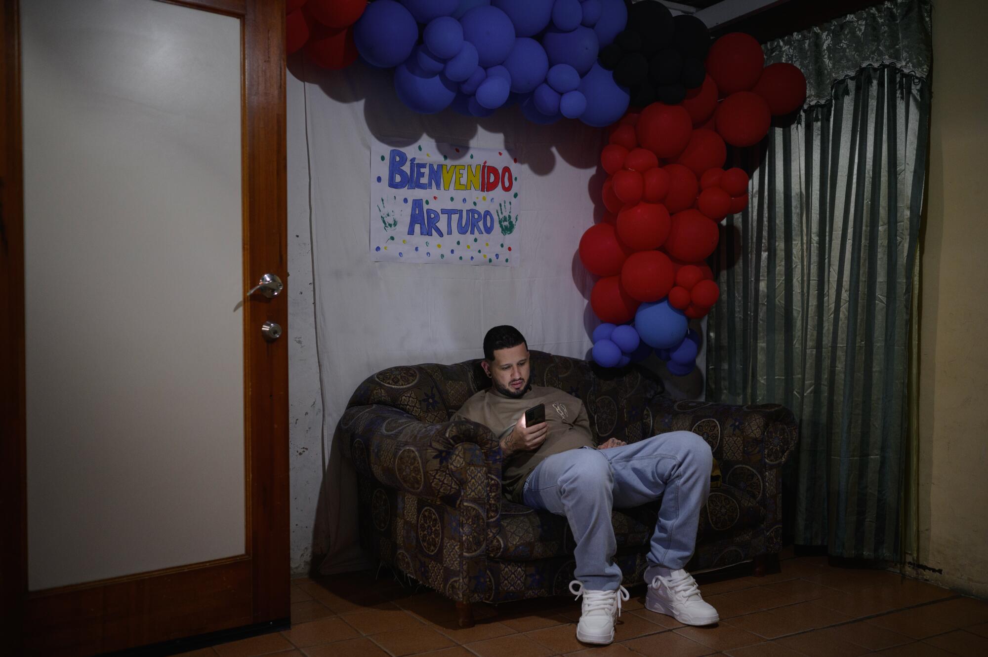 A man with dark hair, seated under a hand-drawn sign and a cluster of red and purple balloons, looks at his phone 