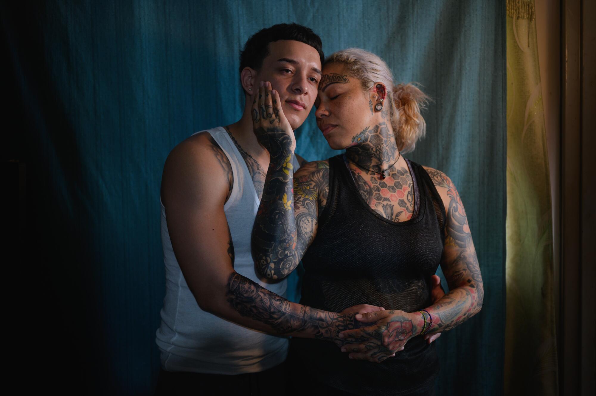 A blond woman covered in tattoos holds the face of a young man, with her other hand over his 