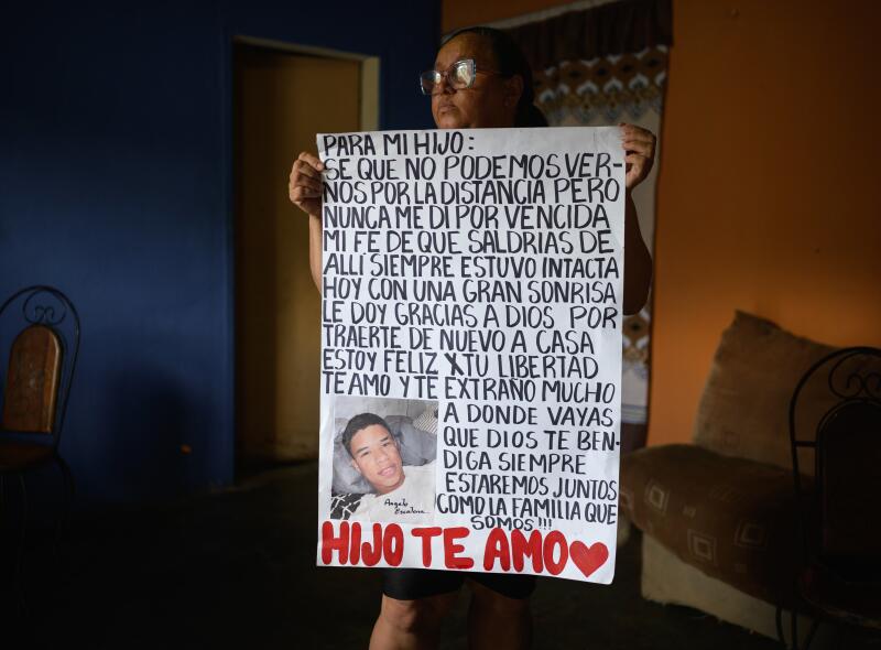 A woman with glasses, holding a large white hand-drawn poster with words and a photo of a young man