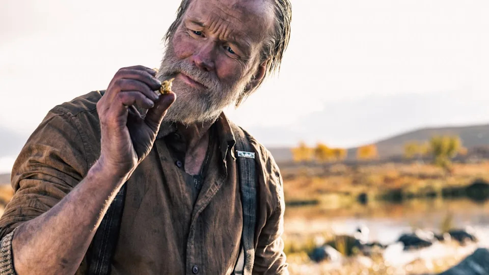 The image shows a man in the outdoors examining a chunk of gold that is held between his thumb and finger.