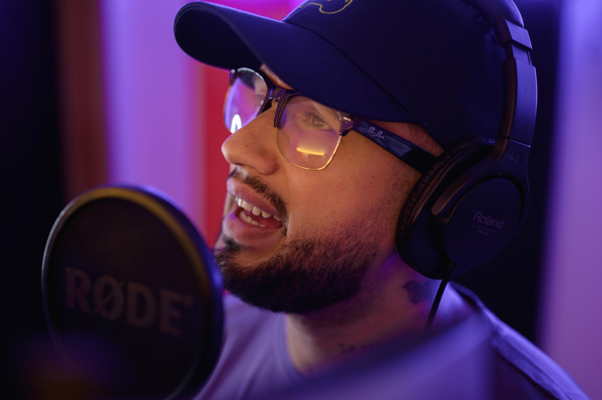 A man with a dark beard, wearing glasses, headphones and a dark ballcap, sings 