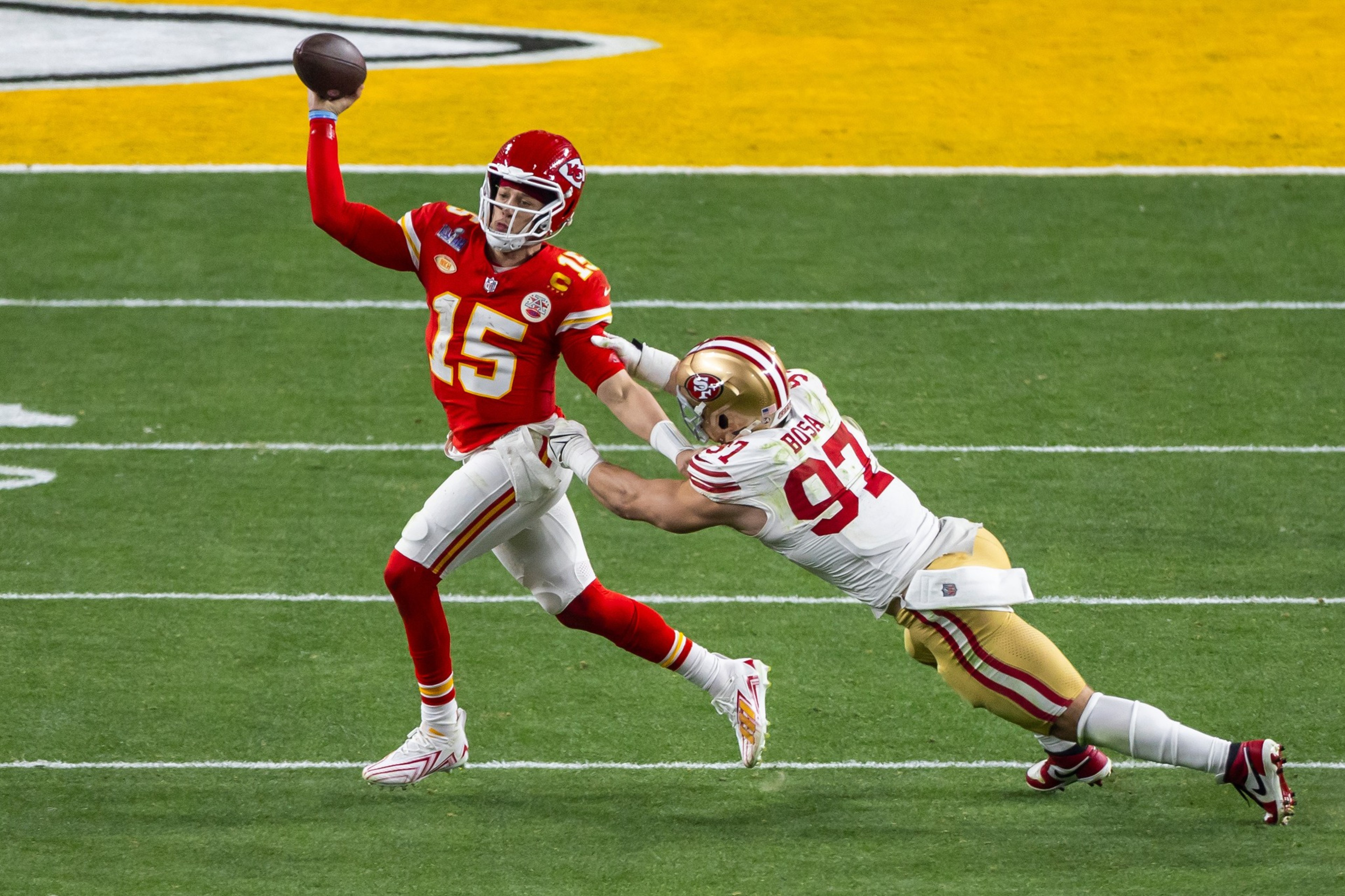 A football player in a red uniform throws a pass while being tackled by an opponent in a white and gold uniform on a green field.
