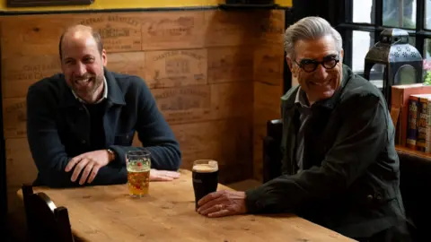 AppleTV+/PA Prince William and Eugene Levy sharing a drink in a pub, both are smiling