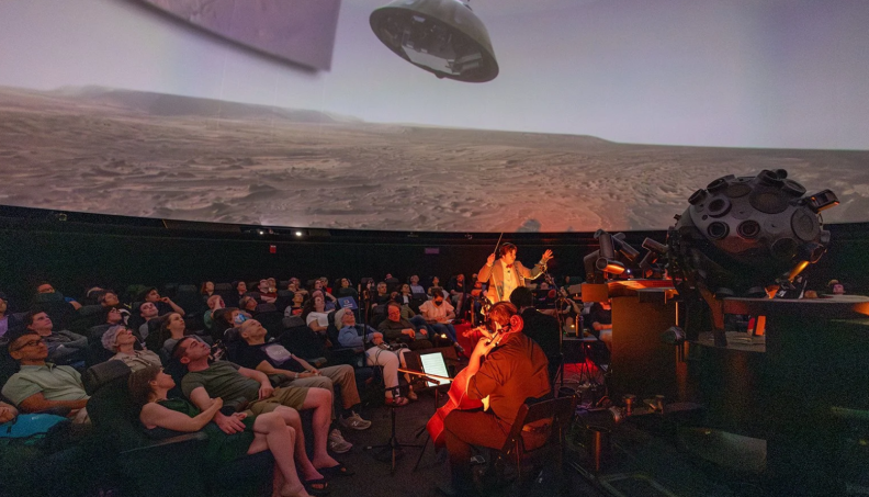 An audience sine in a domed room, watching a projection of a film on the ceiling as a conductor and orchestra play.