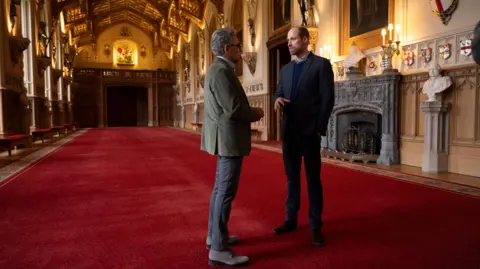 AppleTV+/PA Prince William showing Eugene Levy inside Windsor Castle, they are in a large room or corridor with red carpet and ornate furnishings