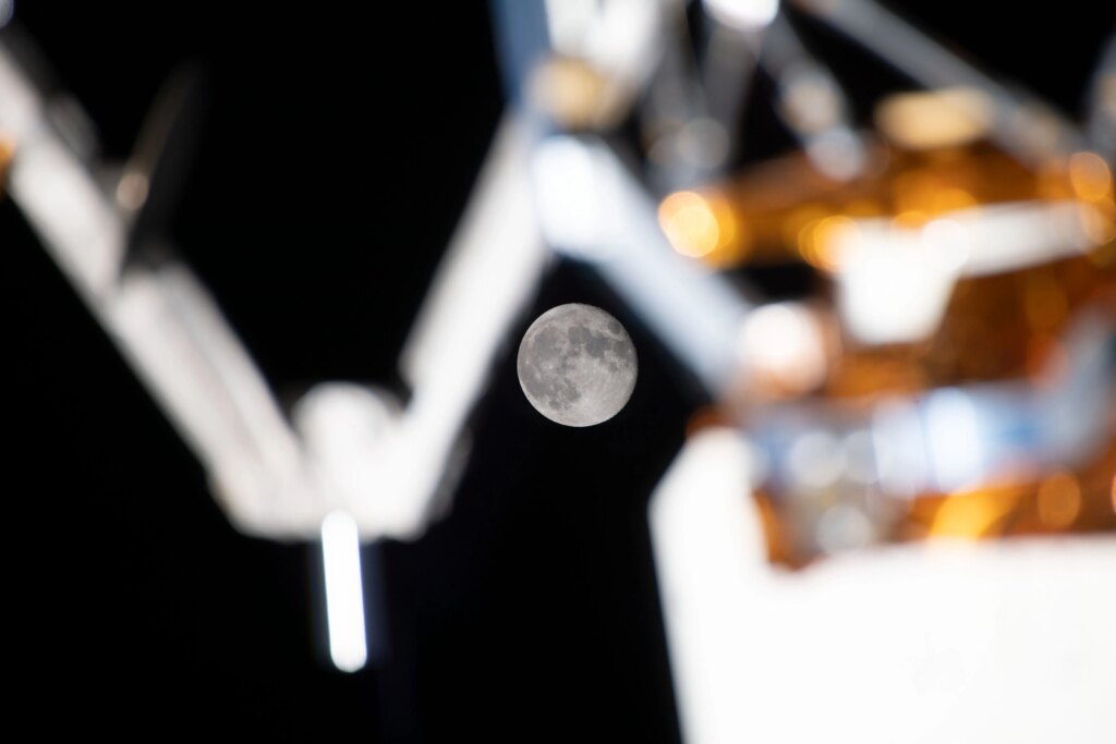 A full Moon is clearly visible in the dark background of space, framed by the blurred mechanical arms and equipment on the International Space Station’s exterior. Caption: The Moon shines between the space station’s external equipment. (Credit: NASA)