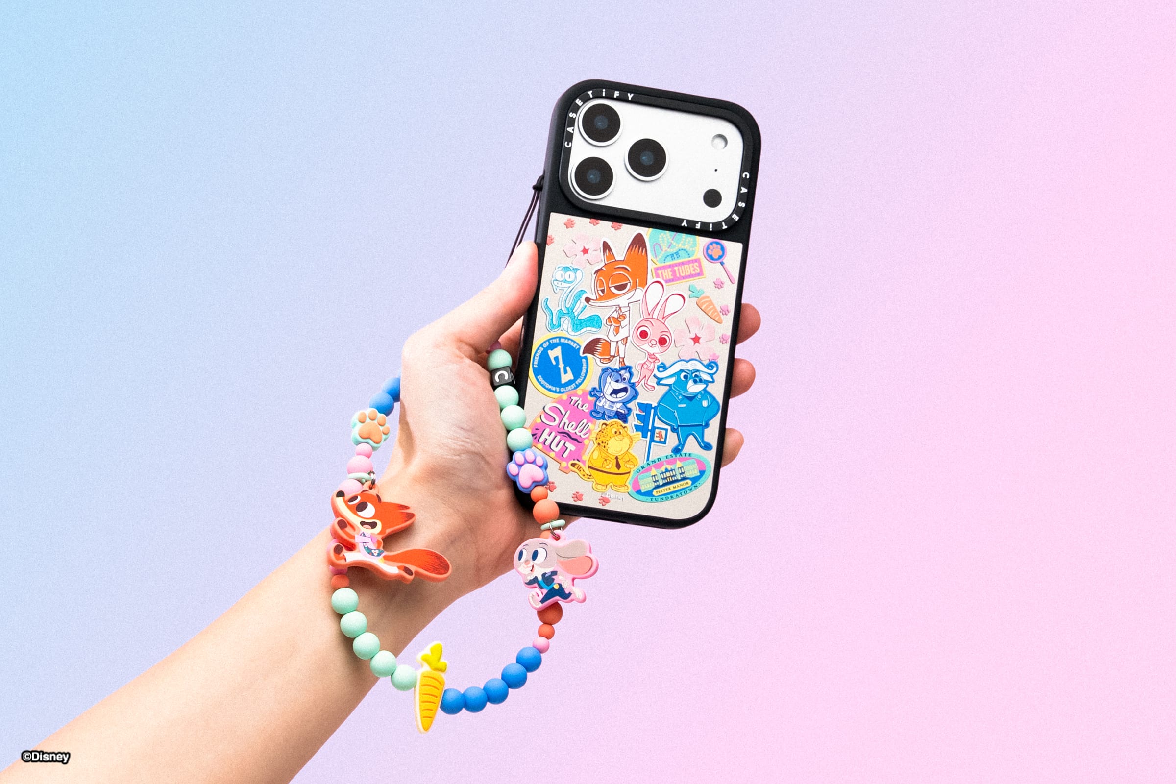 A hand holds a phone decorated with Disney Store stickers and a beaded strap, reminiscent of park souvenirs, on pastel backdrop.