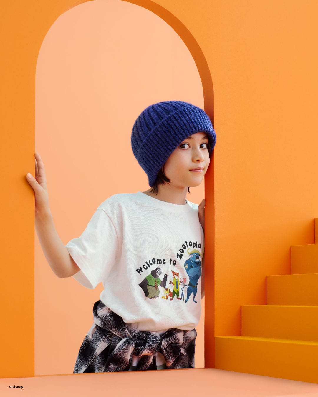 A young person in a blue knit beanie and Zootopia 2 shirt stands by orange stairs in an archway, like park entryways at Disney.