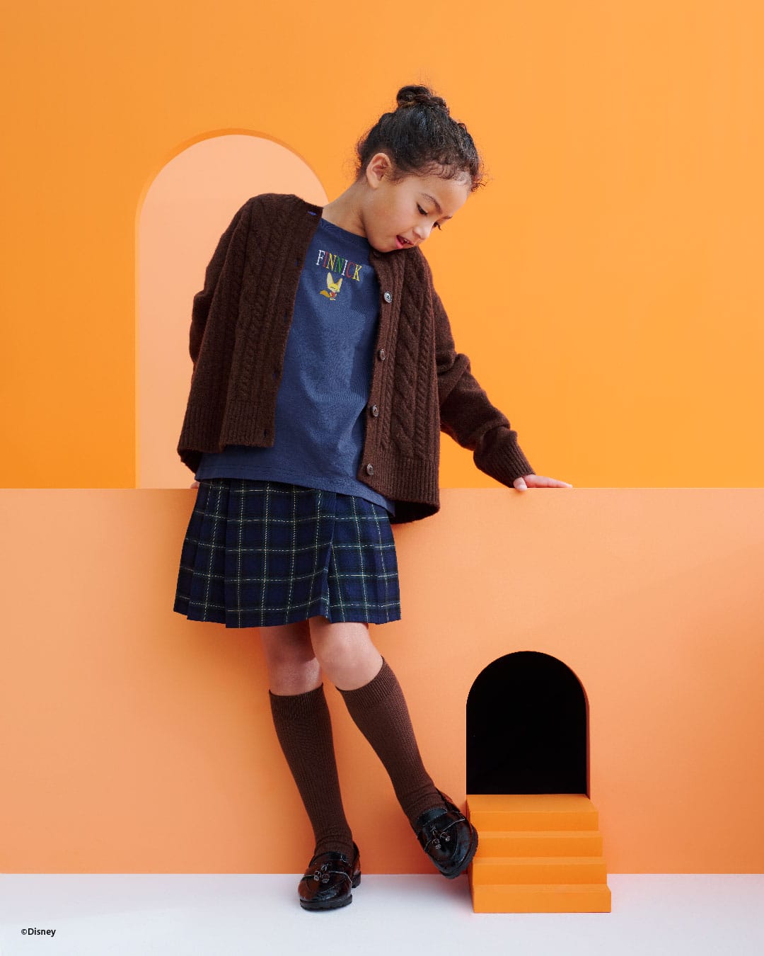 Young girl in a navy plaid skirt and brown cardigan stands by an orange wall, holding Disney Store merchandise, evoking Main Street.