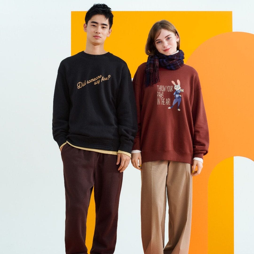 Two people in Disney-themed sweatshirts pose before a bold orange and white backdrop, echoing Disney Parks’ playful style.