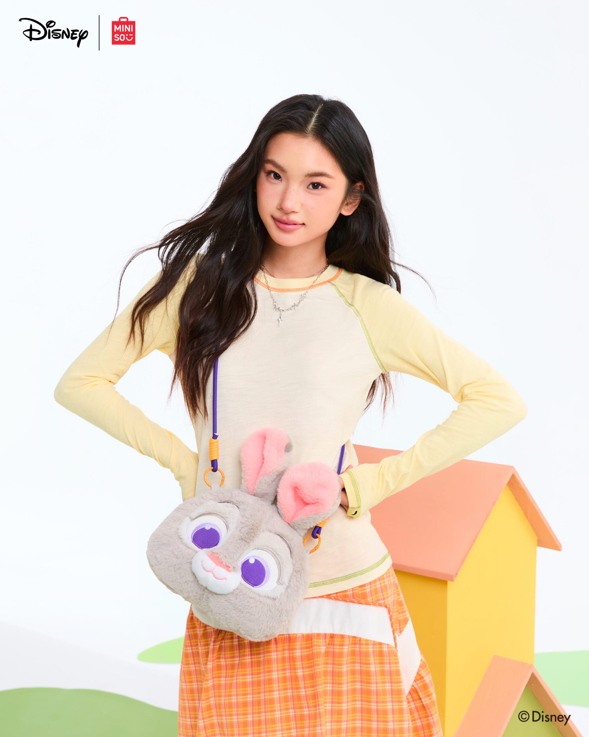 At a Disney park, a young woman holds Disney Store bags, wearing a yellow top, orange plaid skirt, and carrying a fluffy bunny bag.